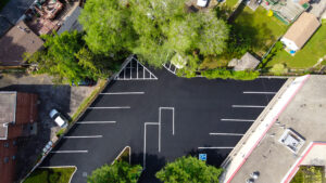 Aerial view of a freshly sealcoated parking lot with deep black asphalt, crisp white parking lines, and clearly marked spaces, surrounded by trees and nearby buildings.