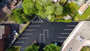 Aerial view of a freshly sealcoated parking lot with deep black asphalt, crisp white parking lines, and clearly marked spaces, surrounded by trees and nearby buildings.