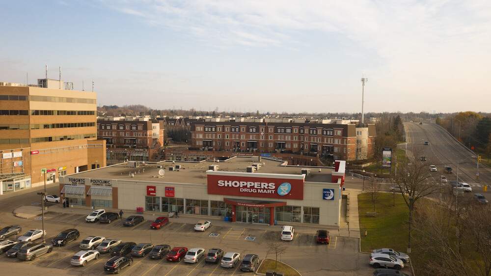 Commercial plaza and asphalt parking lot in Thornhill, Ontario, featuring retail storefronts, parked vehicles, and a major roadway running through the suburban community.