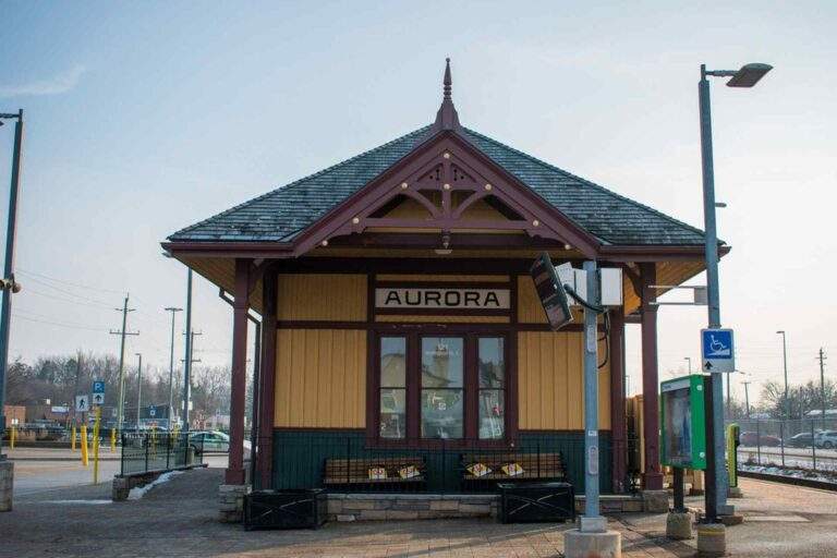 Historic Aurora train station building with a peaked roof, wooden trim, and a central entrance, set beside transit signage and a small platform area.