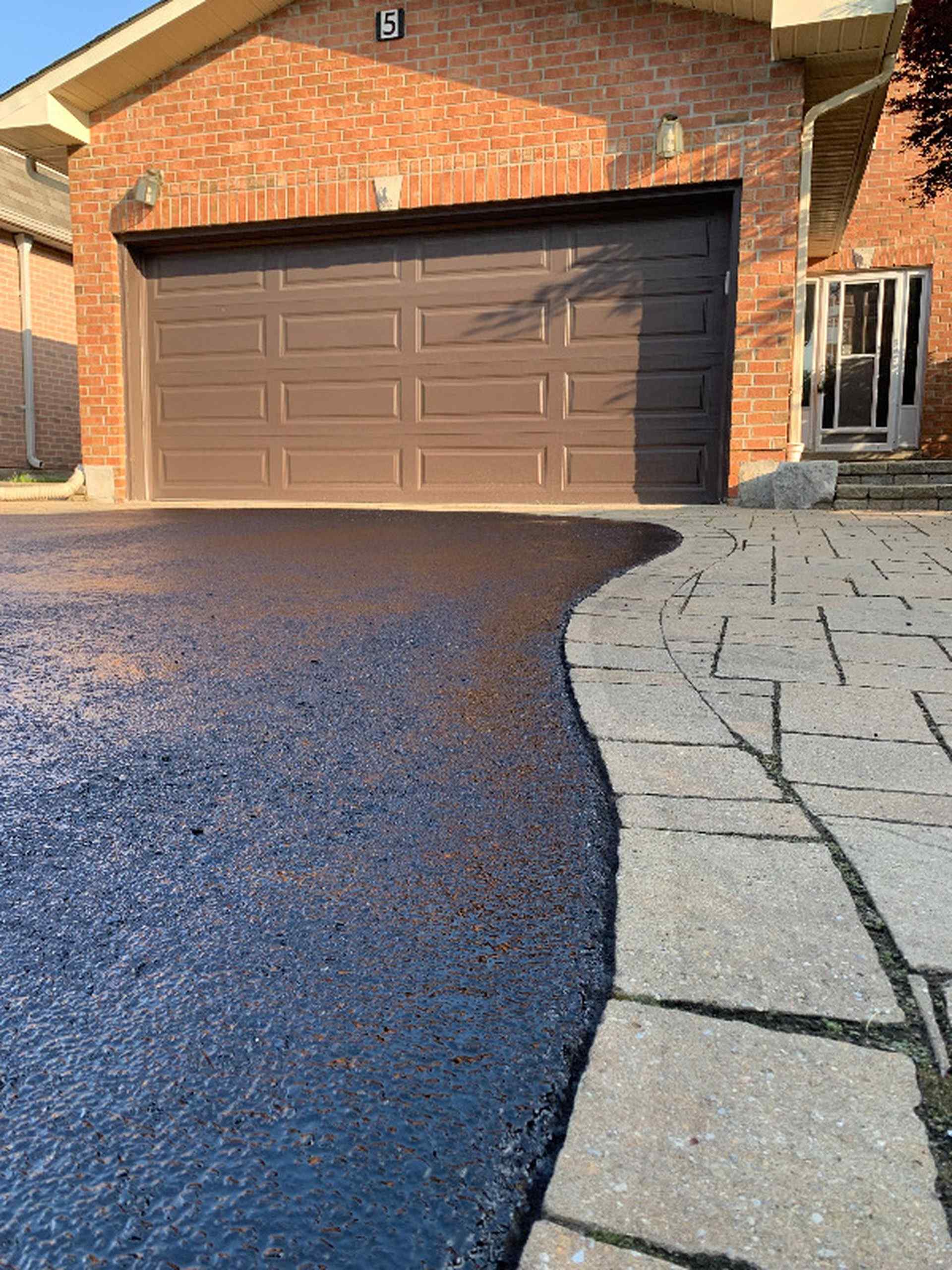 Freshly sealcoated residential asphalt driveway beside a brick home, featuring a smooth black surface and decorative stone walkway leading to a double garage.