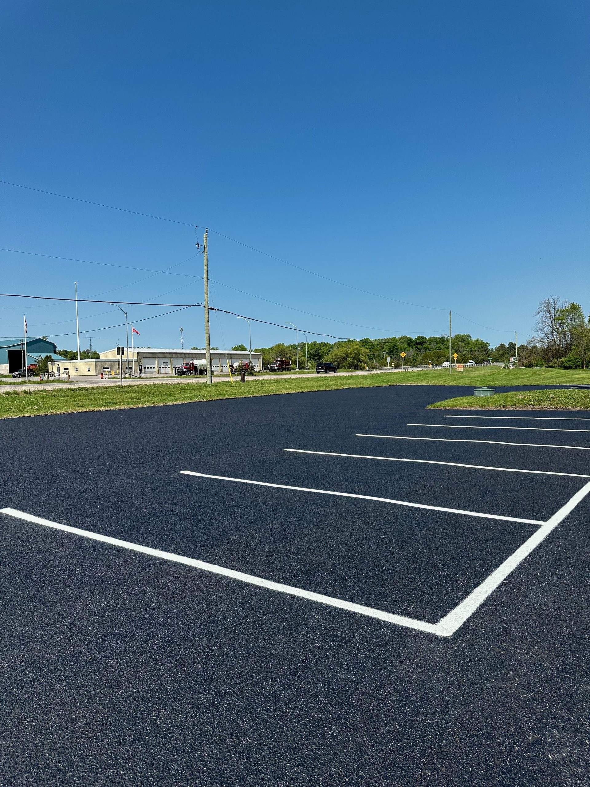 Freshly paved asphalt parking lot with newly painted parking stall lines, showcasing a smooth blacktop surface and clean pavement markings under a clear blue sky.