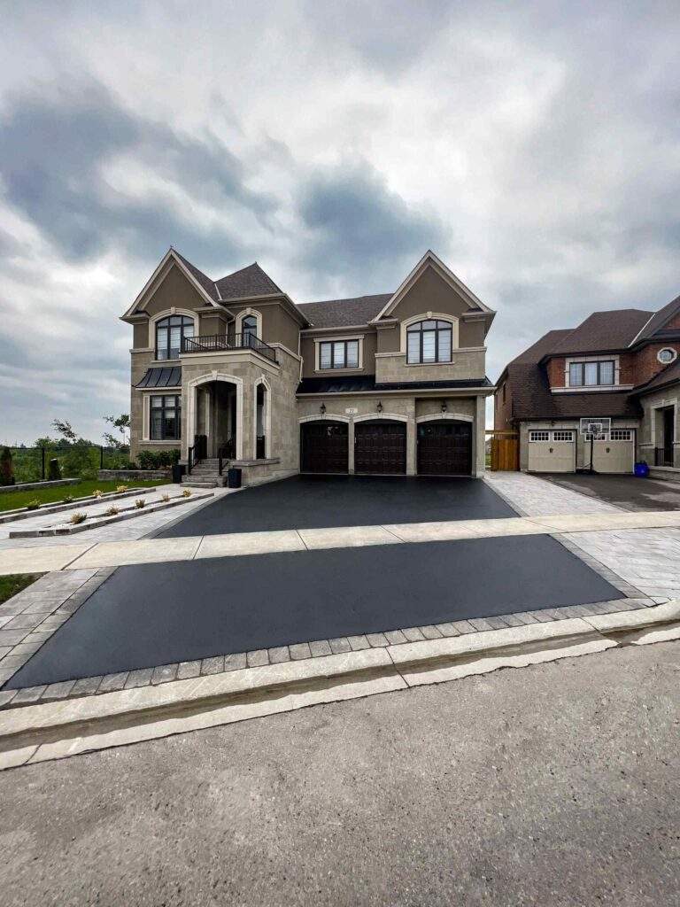 Newly paved residential asphalt driveway in front of a large custom home, featuring smooth black pavement, decorative stone borders, and a three-car garage in an upscale neighborhood.