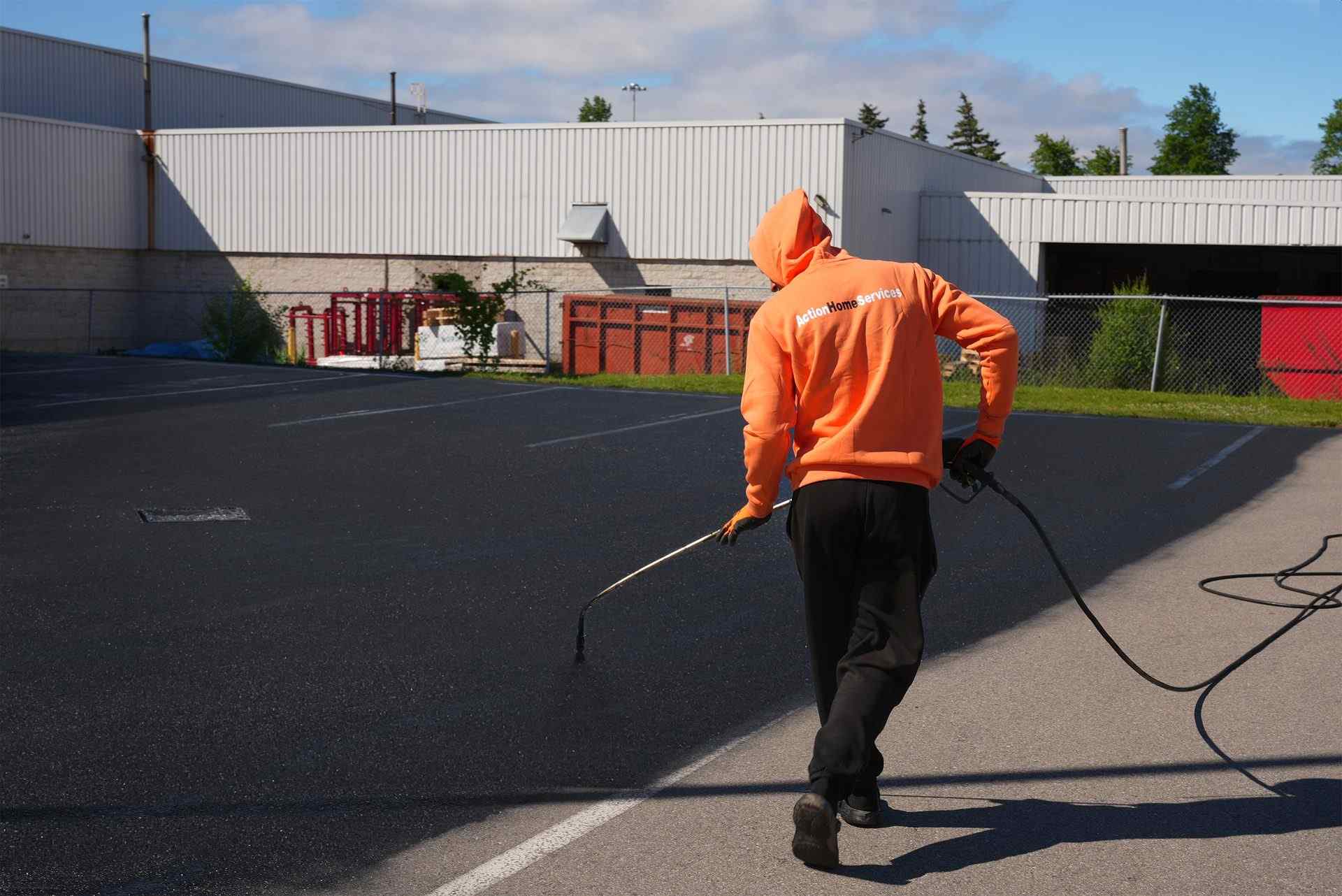 Worker applying asphalt sealcoating to a commercial parking lot, spraying a protective coating across the pavement surface in front of an industrial building to extend asphalt life and improve appearance