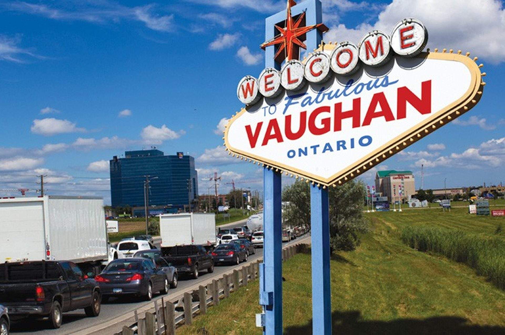 “Welcome to Vaughan, Ontario” roadside sign with traffic-filled highway and city buildings in the background under a blue sky.