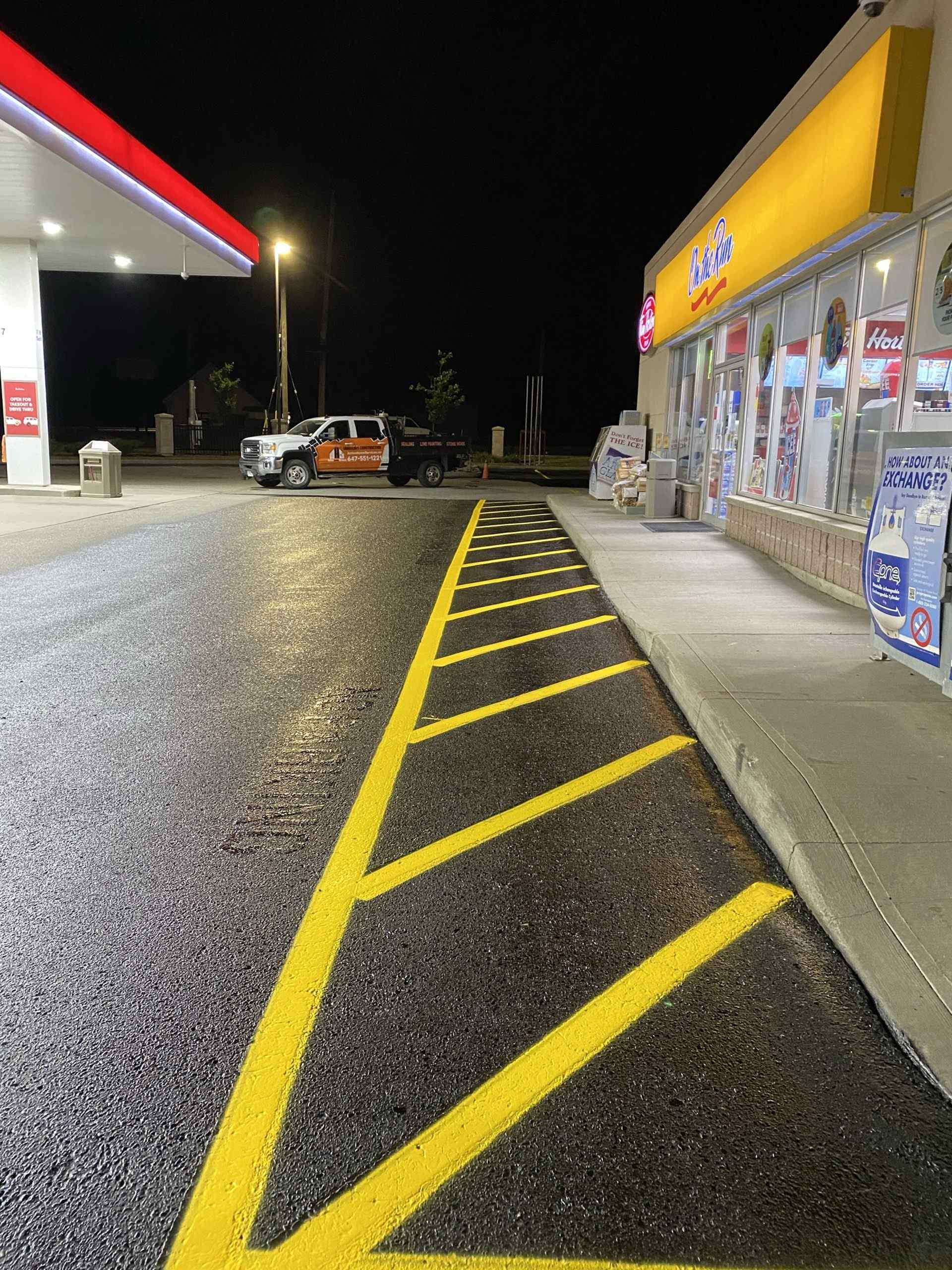 Freshly striped yellow no-parking hatch markings beside a convenience store and gas station at night, highlighting clear pavement markings on a newly paved asphalt surface for improved traffic flow and pedestrian safety.
