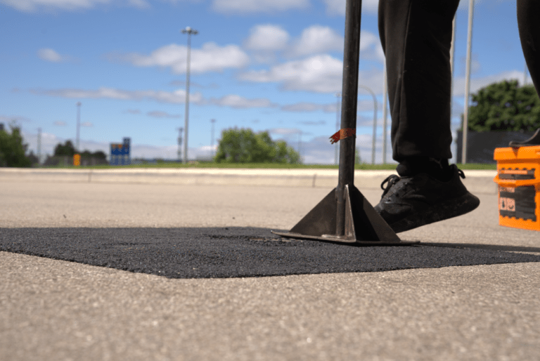 Close-up of a field technician compacting a fresh hot-mix patch using a manual asphalt tamper tool during a commercial property repair